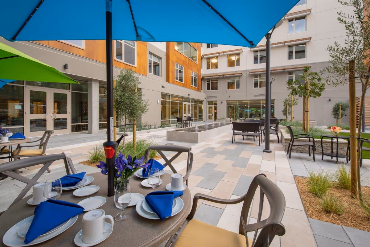 Outdoor courtyard with umbrella-shaded dining tables set with blue napkins and placeware at a senior living facility.