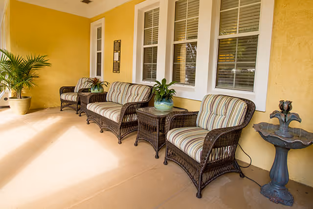 Covered outdoor porch with wicker chairs and small tables and potted plants along a yellow building wall.