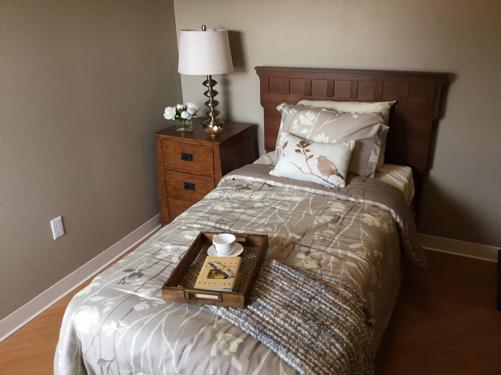A single bed with floral patterned bedding and a decorative pillow featuring a bird design. A wooden nightstand with three drawers is beside the bed, holding a modern lamp and a small vase with white flowers. A wooden tray with a cup, saucer, and a book is placed on the bed. The room has beige walls and wooden flooring.