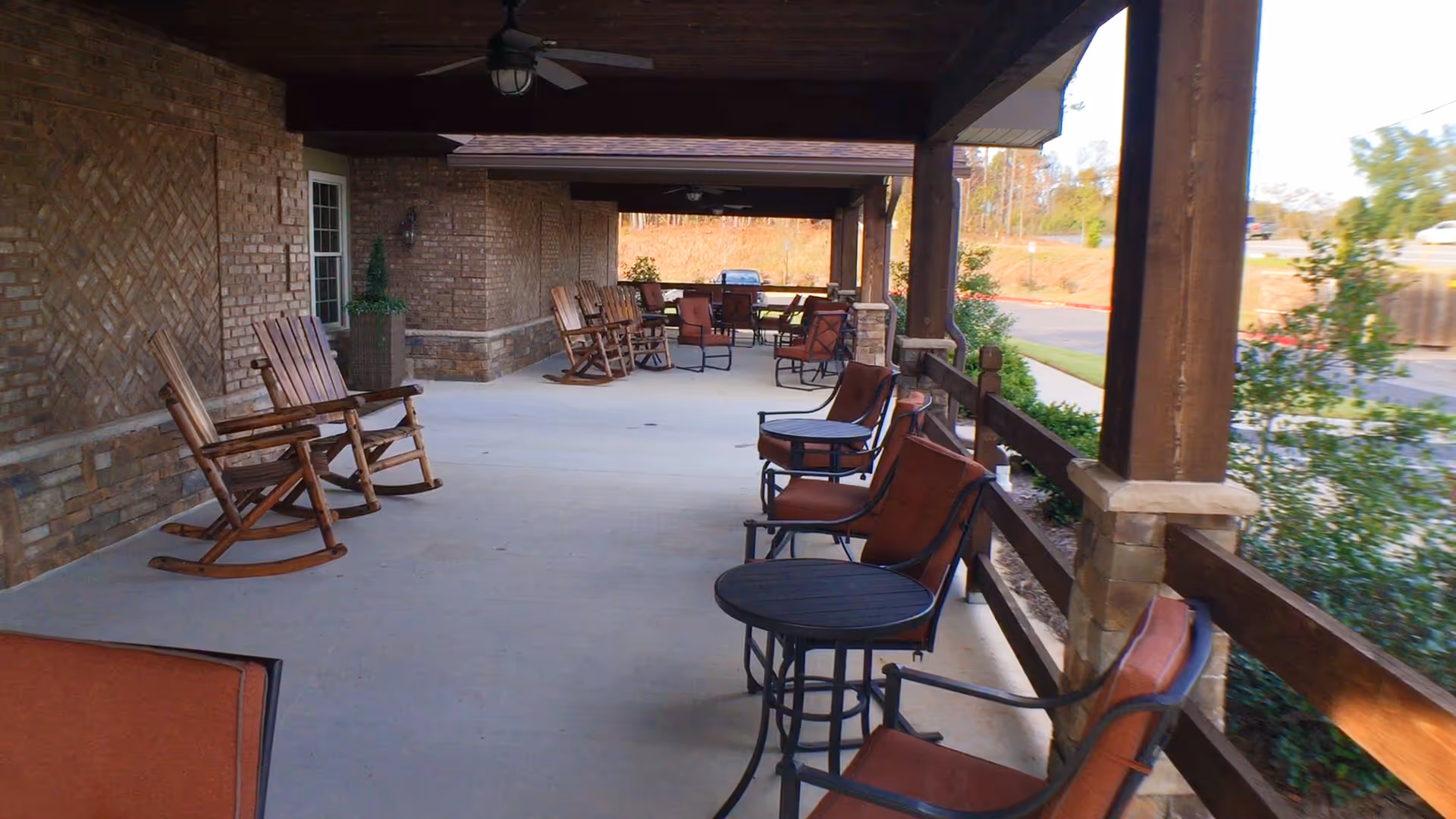 Covered outdoor patio area with wooden rocking chairs and metal chairs with cushions arranged around small round tables. The patio has a brick wall on one side and wooden posts supporting the roof. There is greenery and a parking area visible beyond the patio.