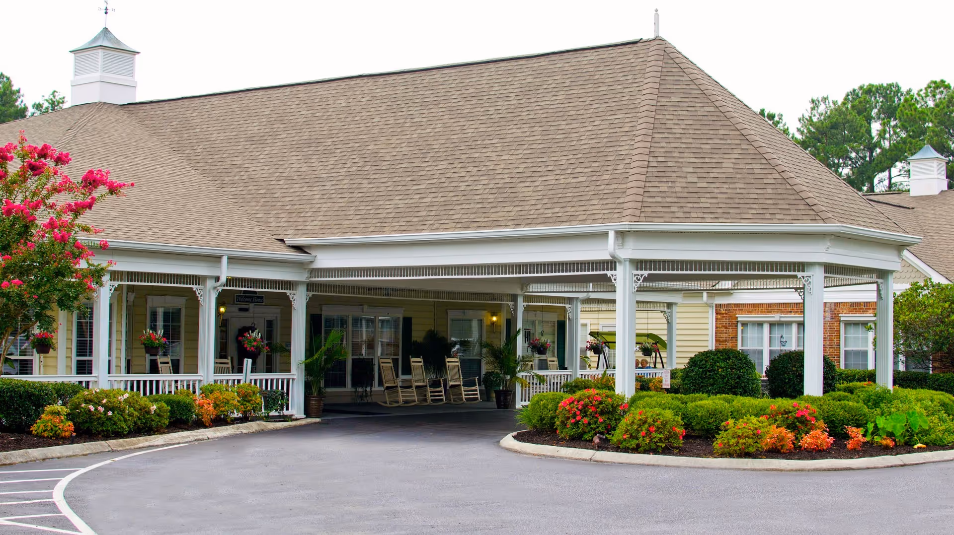 Exterior view of a senior living facility entrance with a covered driveway, white pillars, rocking chairs on the porch, and well-maintained landscaping including bushes and flowering plants.
