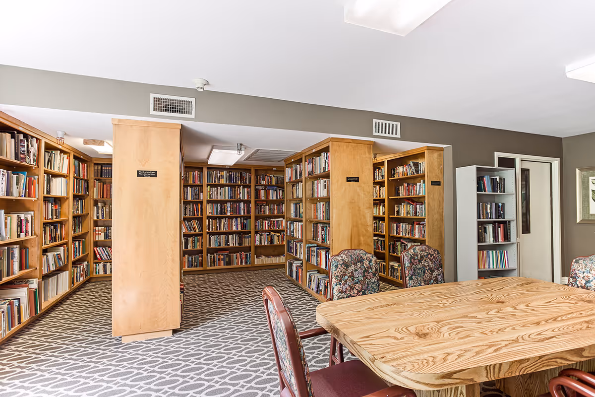 Interior view of a library room with wooden bookshelves filled with books lining the walls and a wooden table surrounded by floral upholstered chairs in the foreground. The room has a patterned carpet and neutral-colored walls with ceiling lights.