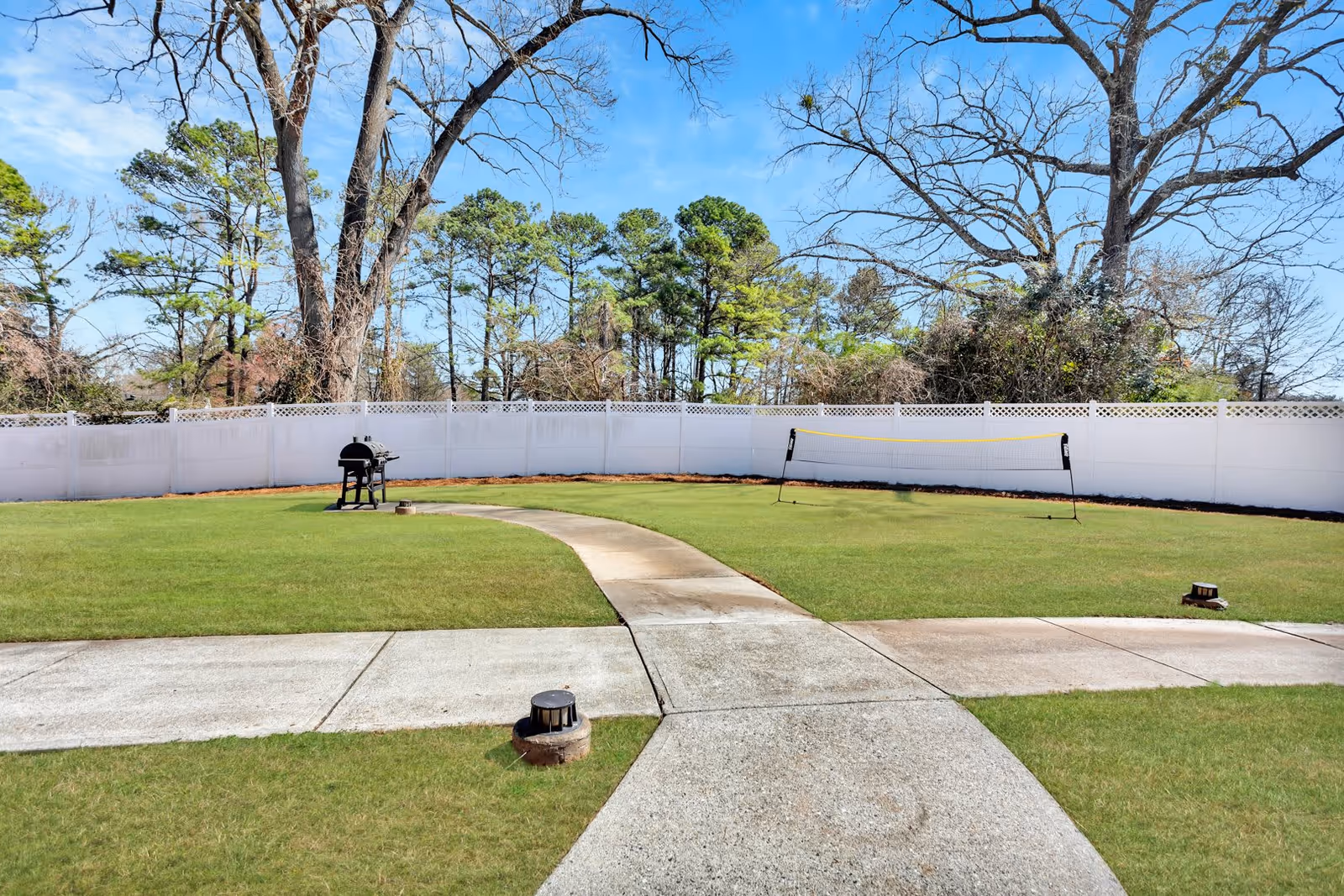 A fenced outdoor area with a green lawn, a concrete pathway, a barbecue grill, and a badminton net set up. Tall trees and a clear blue sky are visible in the background.