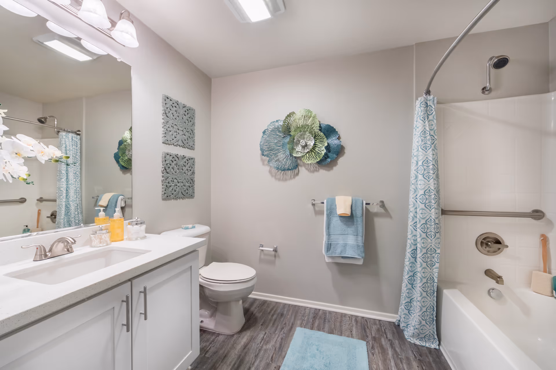 A clean and modern bathroom featuring a white vanity with a sink and faucet, a large mirror with overhead lights, a toilet, and a bathtub with a showerhead and a blue patterned shower curtain. The walls are decorated with blue and green metal wall art and two square carved wooden panels. There are blue and beige towels hanging on a towel rack and a blue bath mat on the wood-look floor.