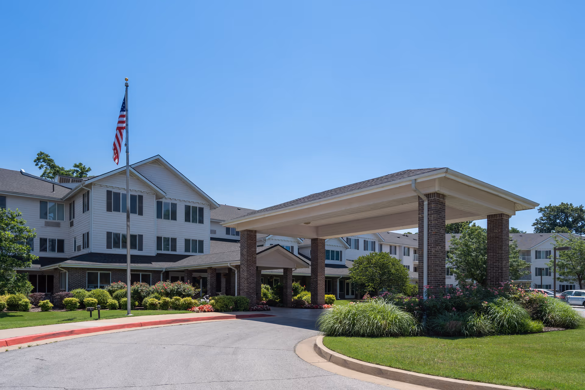 Exterior view of a multi-story senior living facility with a covered entrance supported by brick columns, landscaped greenery, and an American flag on a flagpole under a clear blue sky.