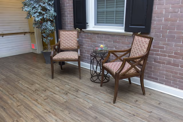 Two wooden chairs with patterned cushions placed against a brick wall under a window with black shutters. A small round glass table with a decorative item is positioned between the chairs. The floor is wooden, and there is a potted plant in the corner near a white paneled wall.