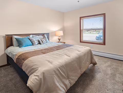 A neatly made bed with a beige and brown comforter and several decorative pillows in a room with beige walls, a window with blinds, a nightstand with a lamp, and carpeted floor.