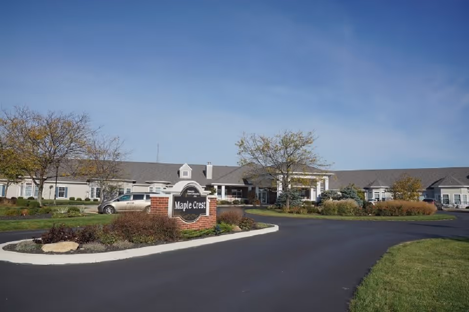 Exterior view of Maple Crest Senior Living Village showing a single-story building with a gray roof, surrounded by landscaped greenery and trees under a clear blue sky. A brick sign with the name 'Maple Crest' is prominently displayed near the entrance driveway.