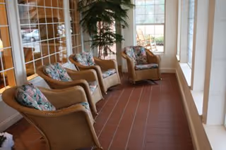 Sunlit sitting area with four wicker armchairs with floral cushions along a windowed wall and a tiled floor.