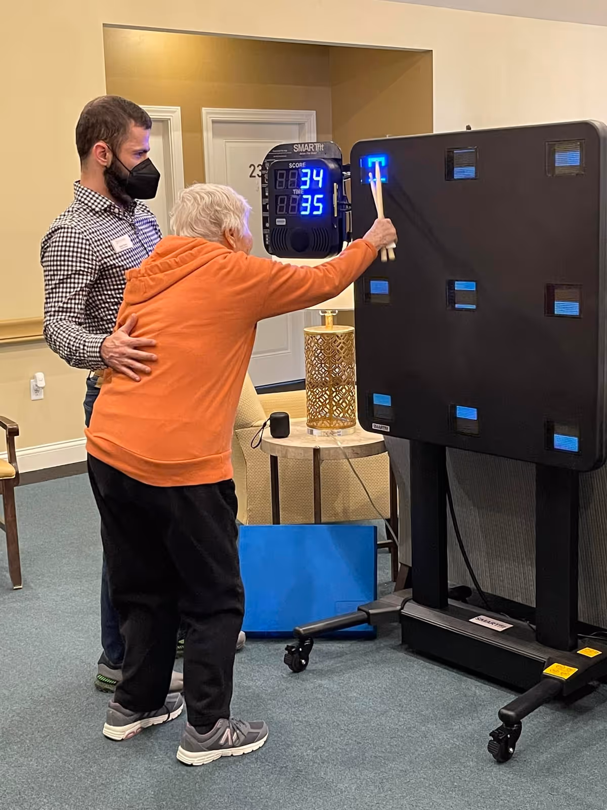An elderly woman wearing an orange hoodie and black pants is playing a reaction game using drumsticks on a black electronic game board with illuminated blue squares. A man wearing a checkered shirt, dark pants, and a black face mask is standing beside her, providing support. The setting appears to be an indoor common area with beige walls, a small table with a decorative lamp, and a door labeled 23 in the background.