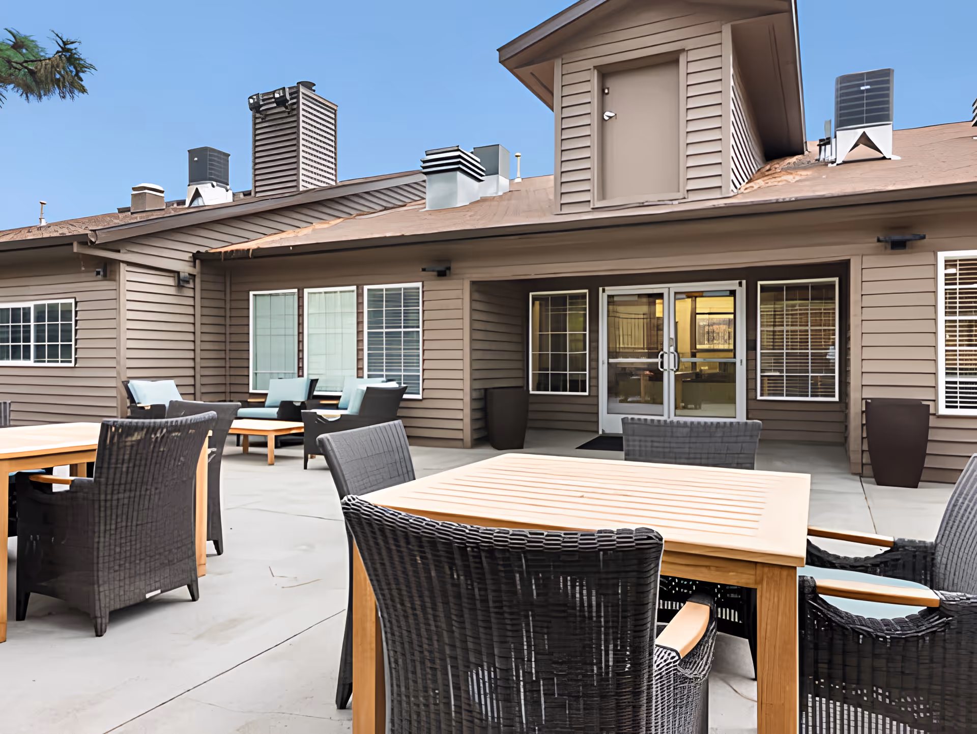 Outdoor patio with wicker chairs and wooden tables in front of a brown memory care building.