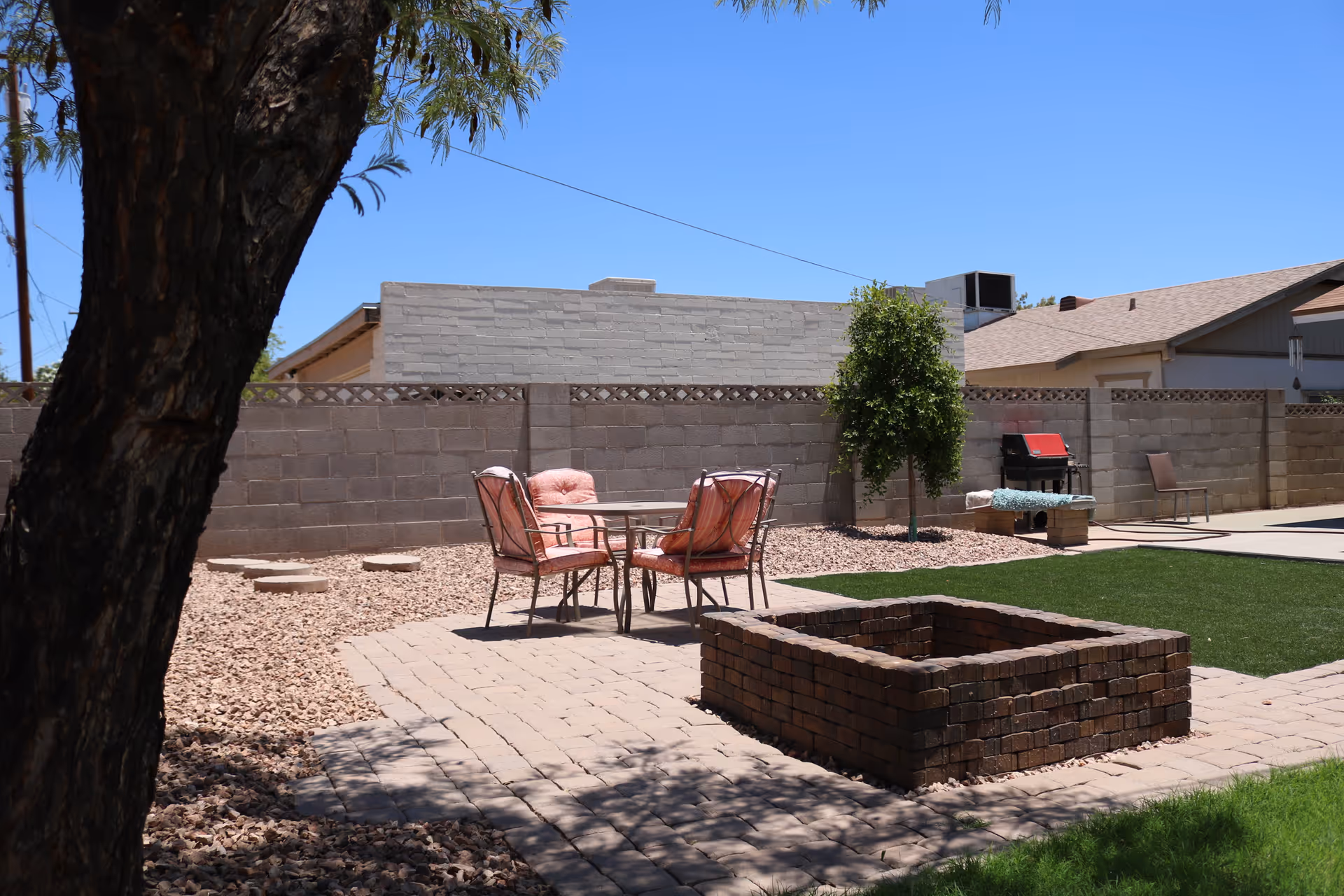 Backyard patio with a table and pink-cushioned chairs, a brick fire pit, artificial turf, and a tree next to a cinderblock wall.