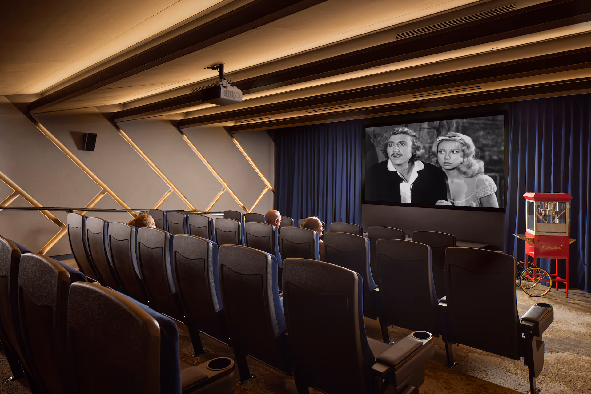 A small movie theater room with rows of cushioned seats facing a large screen showing a black and white film. There are a few people seated watching the movie. The walls have a modern geometric design with soft lighting, and a red popcorn machine is visible on the right side near the screen.