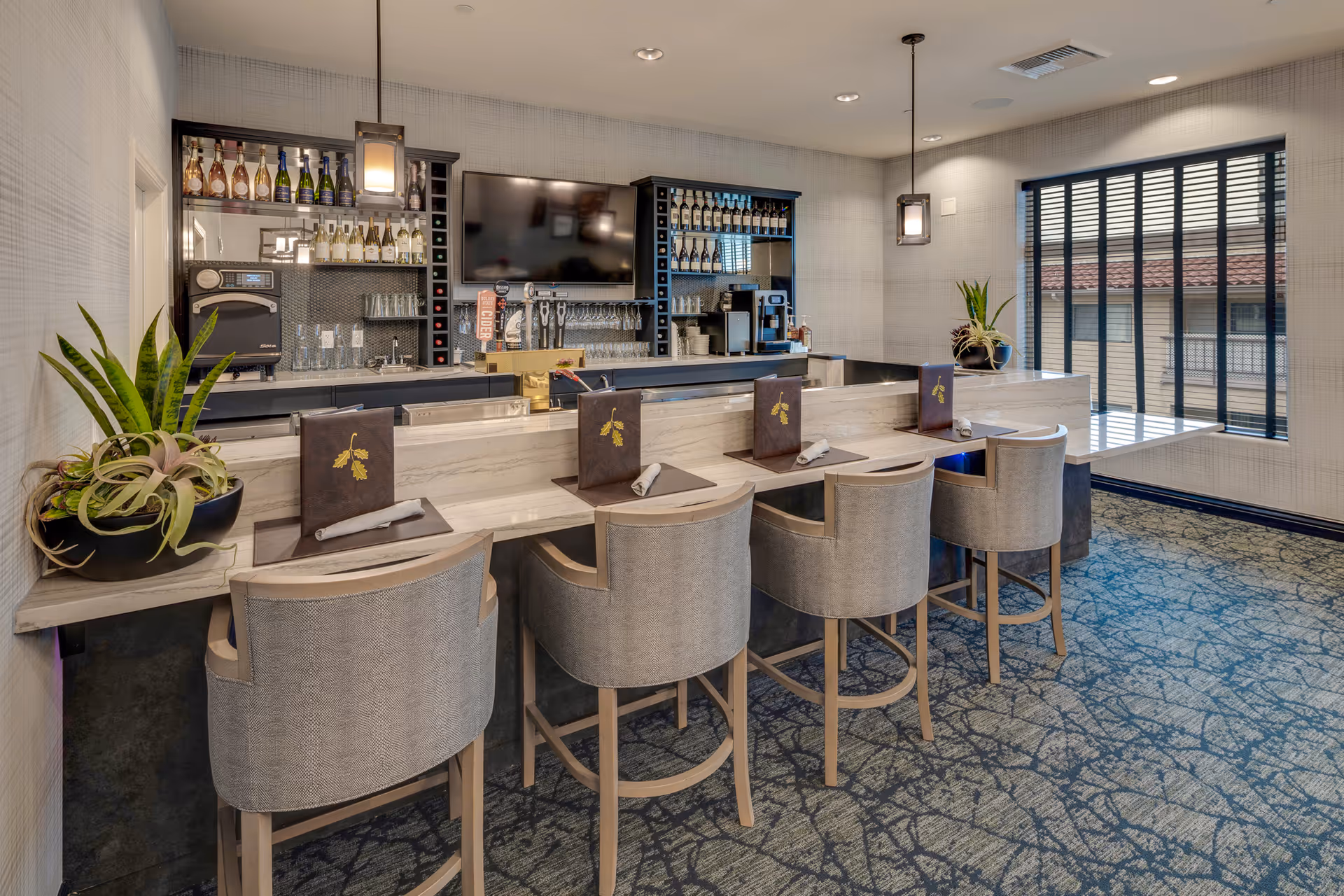 Interior view of a modern bar area with a marble countertop, four upholstered bar stools, menus and napkins on the counter, shelves stocked with wine bottles and glasses, a large flat-screen TV mounted on the wall, and two pendant lights hanging from the ceiling. There are two potted plants on the counter and a large window with black vertical blinds letting in natural light.