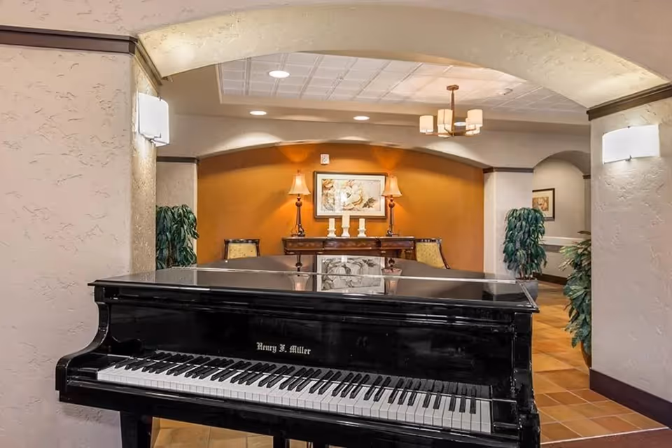 A black grand piano sits in a warmly lit communal lounge with an arched entry, side table, lamps, and potted plants.