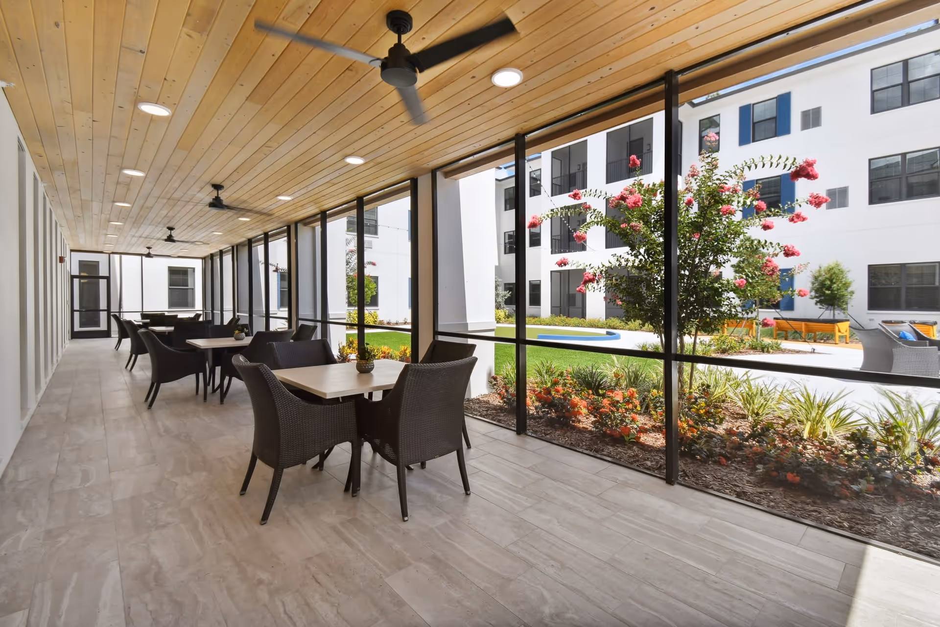 A covered outdoor patio area with multiple tables and wicker chairs, ceiling fans, and recessed lighting. The patio overlooks a landscaped courtyard with flowering bushes and a white multi-story building with blue shutters in the background.