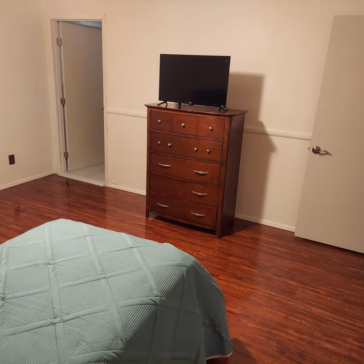 A bedroom with wooden flooring, a bed covered with a light blue quilt in the foreground, a wooden dresser with multiple drawers against the wall, and a flat-screen TV on top of the dresser. There is an open door to a bathroom on the left and a closed door on the right.