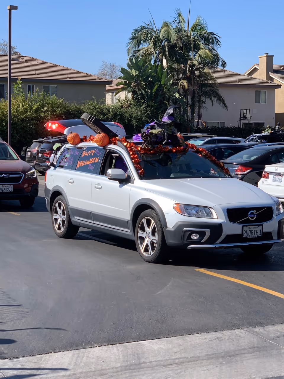 A silver Volvo SUV decorated for Halloween with orange garlands, pumpkins, and a witch hat on the roof. The car is parked in a parking lot with other vehicles and residential buildings in the background. The side window has a message that reads 'Happy Halloween Day!'.