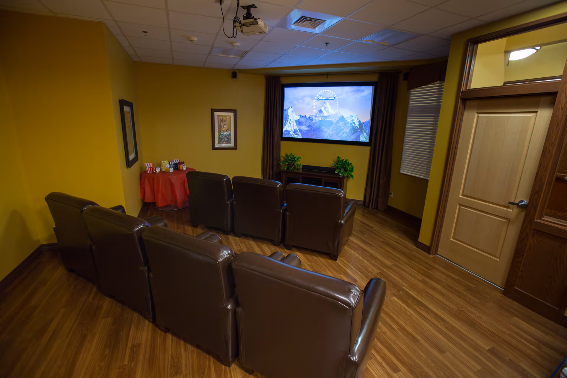 A small home theater room with six brown leather recliners arranged in two rows facing a large screen mounted on a yellow wall. There is a small table covered with a red cloth in the corner holding popcorn and drinks. The room has wooden flooring, a closed wooden door on the right, and framed pictures on the walls.