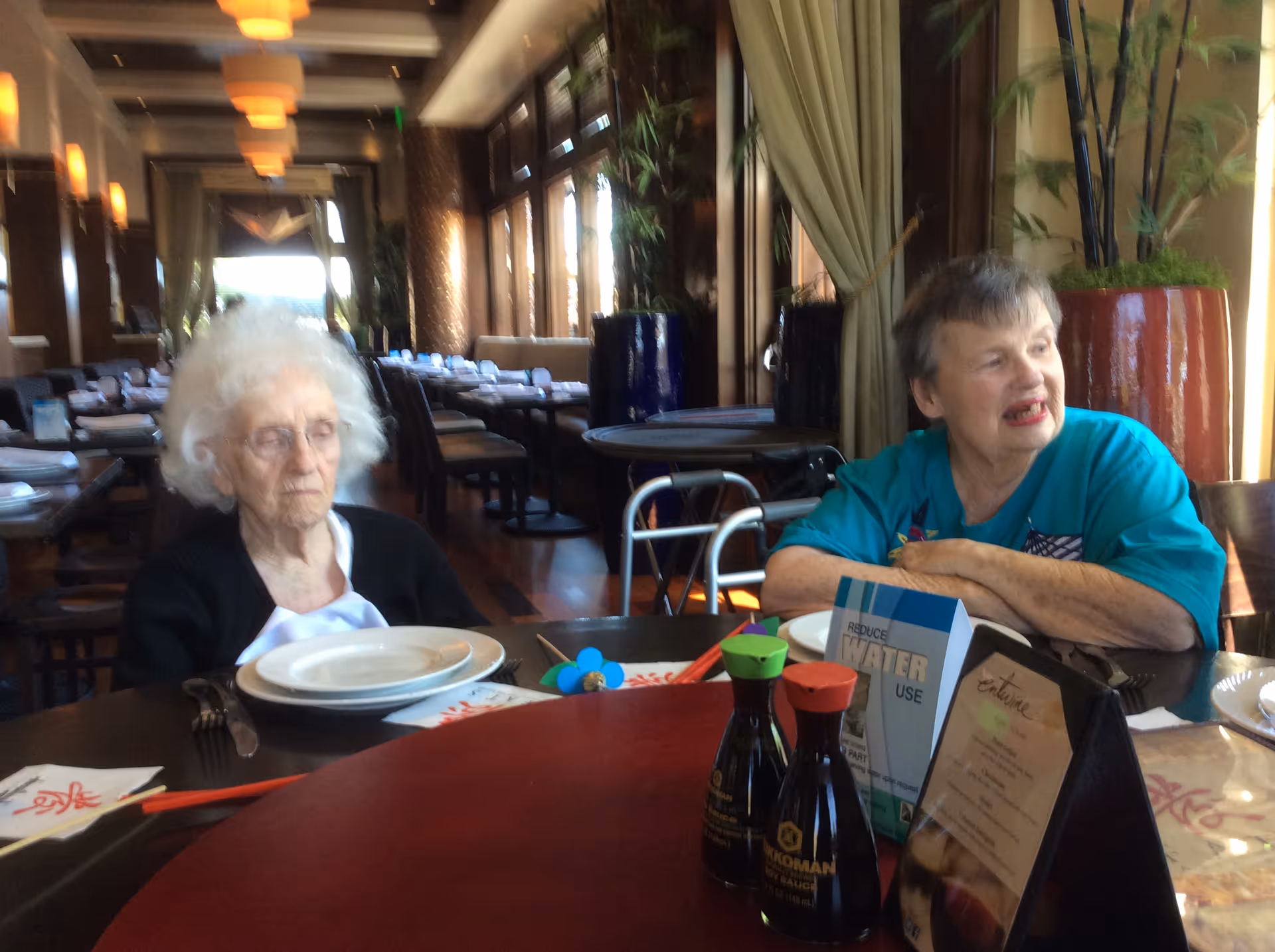 Two elderly women sitting at a round dining table in a restaurant or dining area with plates and condiments on the table. The background shows multiple tables and chairs, large windows with curtains, and warm lighting fixtures.