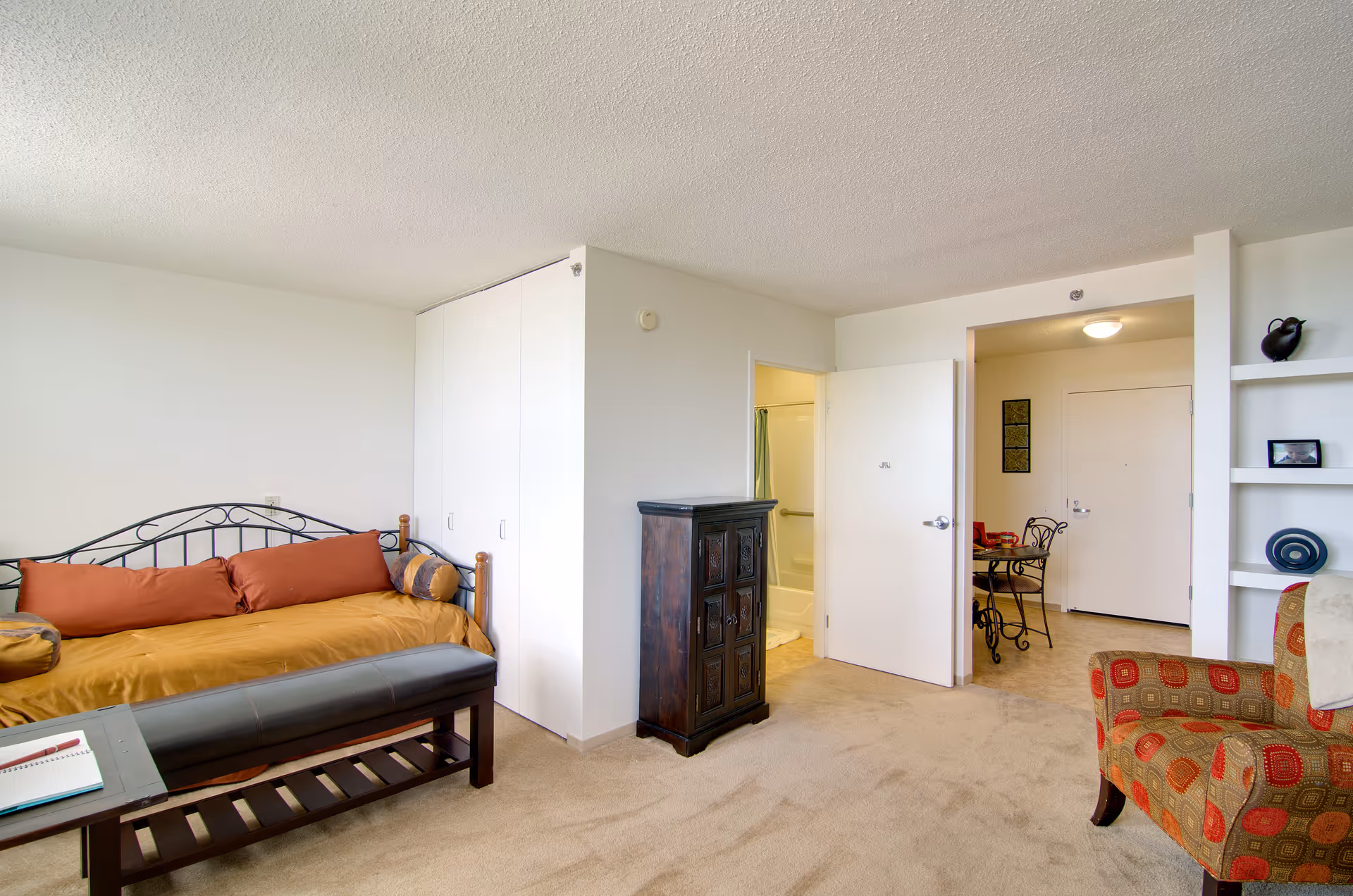 A bright and clean senior living room with a daybed featuring orange and brown cushions, a dark wooden bench at the foot of the bed, a patterned armchair, and a small dark wooden cabinet. The room has beige carpet and white walls. An open door reveals a bathroom with a shower curtain, and another open doorway leads to a small dining area with a round table and chairs near the entrance door. Built-in white shelves hold decorative items.