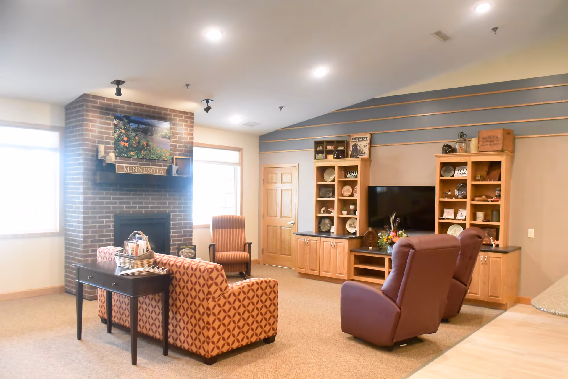 A cozy living room with a patterned orange sofa, two brown recliners, and a striped armchair. There is a brick fireplace with a photo of flowers and a sign that says 'MINNESOTA' above it. Two large windows flank the fireplace. On the right wall, there is a wooden entertainment center with shelves holding decorative items and a flat-screen TV. The room has beige carpet and recessed ceiling lights.