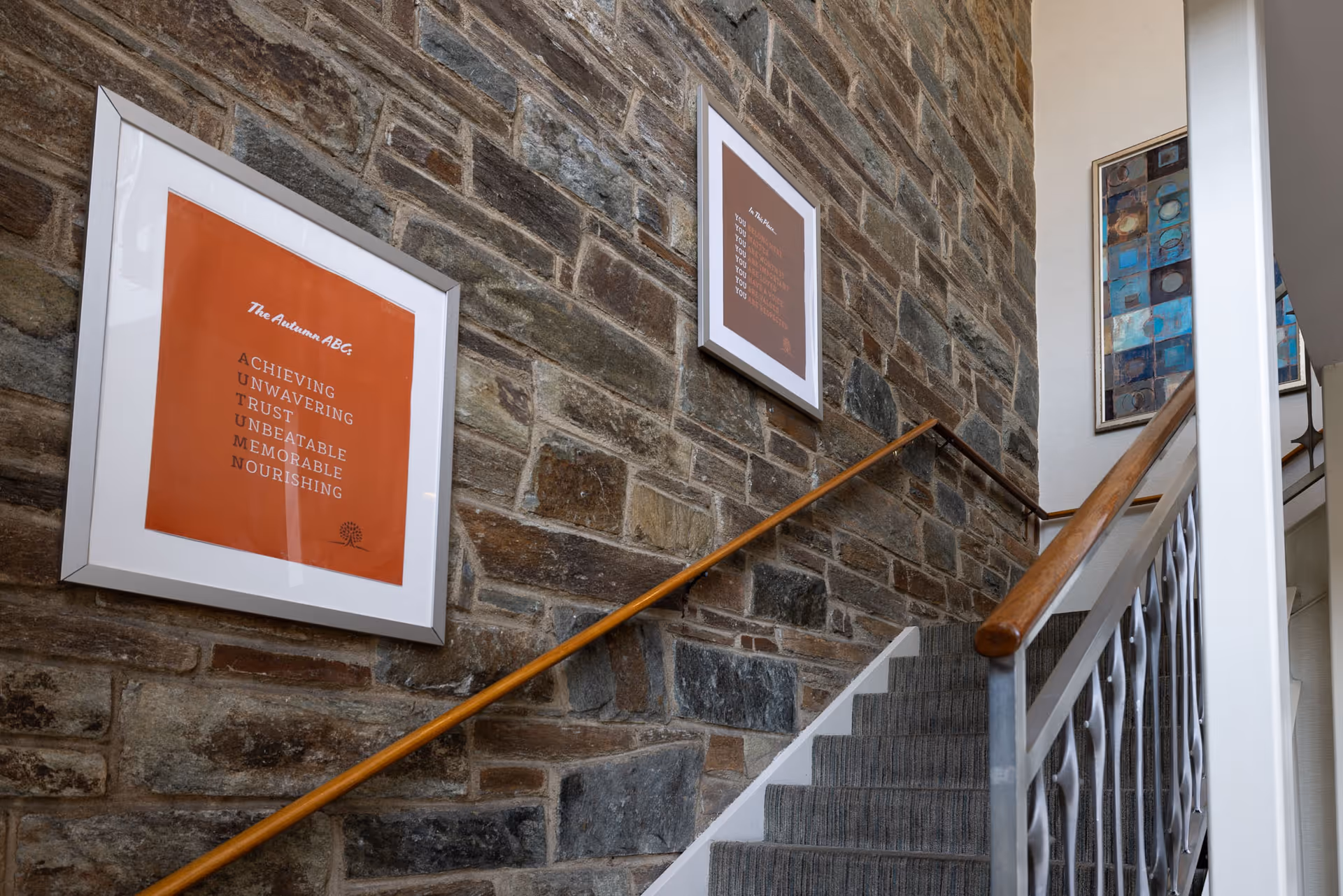 Interior view of a staircase with a stone wall on one side. Two framed artworks with orange backgrounds and white text are hung on the stone wall. The staircase has a wooden handrail and a metal railing with a decorative pattern. A third framed artwork with abstract blue and brown patterns is visible on the adjacent white wall.