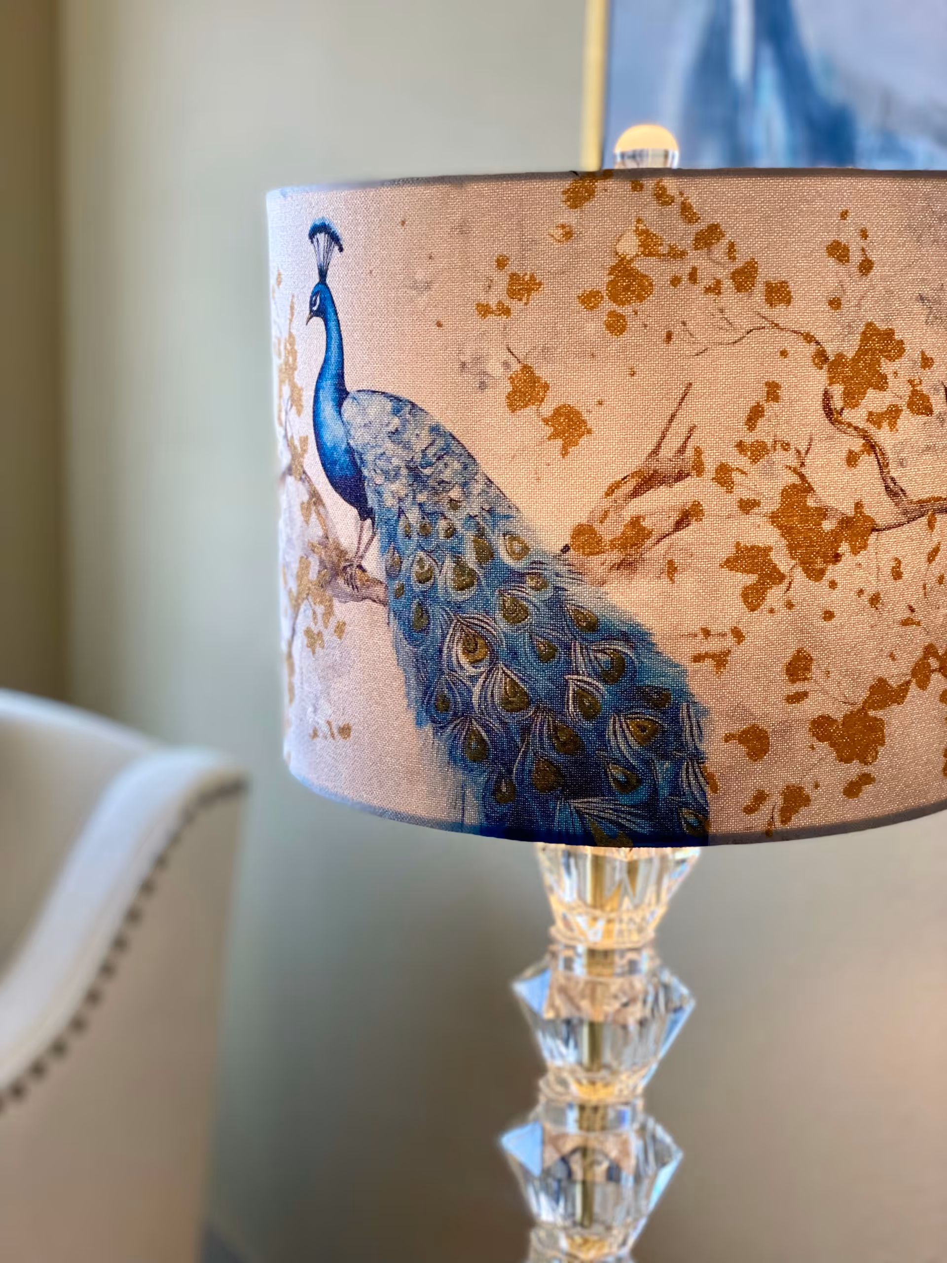 Close-up of a decorative lamp with a lampshade featuring a painted blue peacock and gold floral accents. The lamp base is made of clear, faceted crystal. A beige upholstered chair with nailhead trim is partially visible in the background.