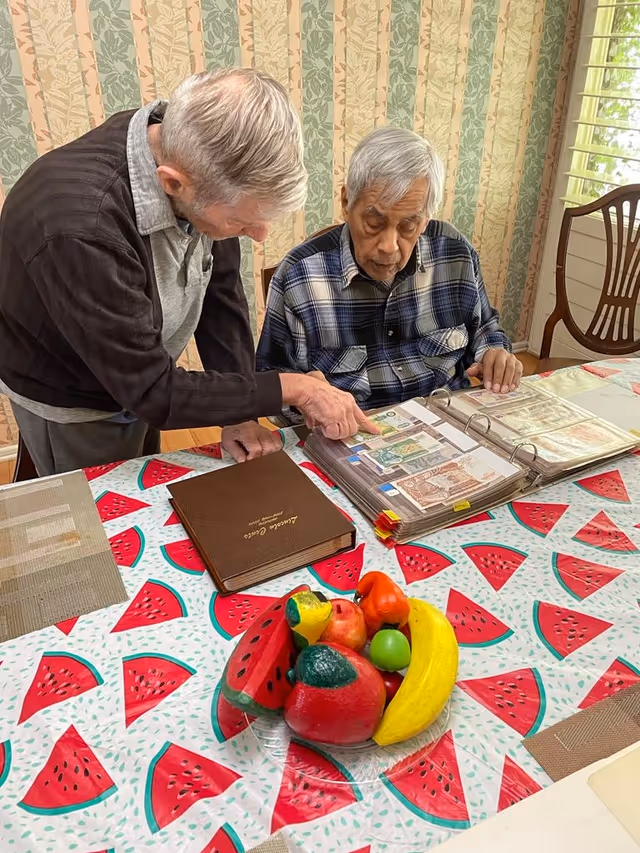 Two elderly men sitting at a table covered with a watermelon-patterned tablecloth. One man is seated and looking at a binder with currency notes, while the other man is standing and pointing at the binder. There is a bowl of artificial fruit on the table and a wooden chair near a window with blinds in the background.