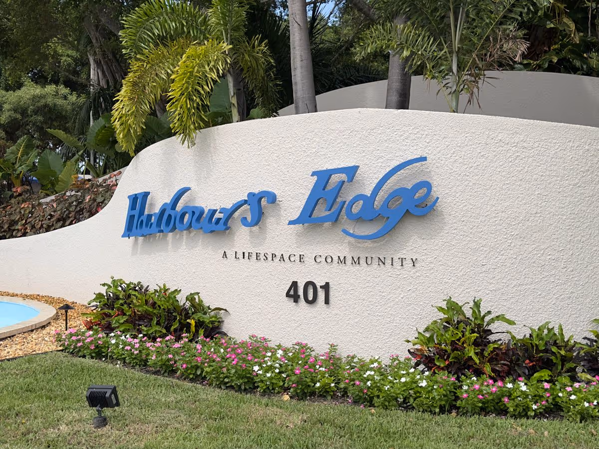 Curved white entrance wall with blue "Harbour's Edge" sign, the words "A Lifespace Community", address number 401, and landscaped plants in front.