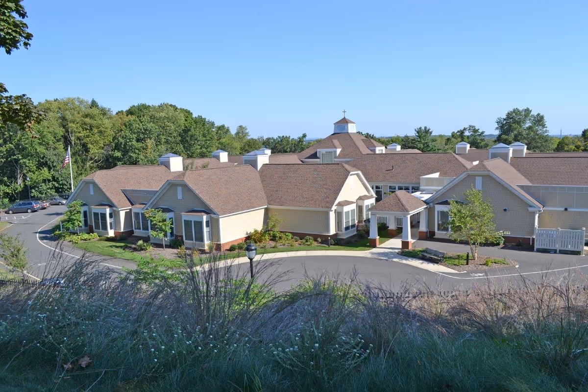 Exterior view of a single-story senior living facility building with beige siding and brown roofs, surrounded by greenery and a clear blue sky.
