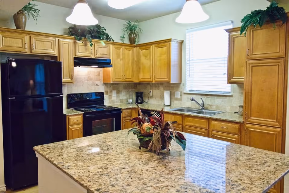 A kitchen with wooden cabinets, a black refrigerator, a black stove with a range hood, a double sink under a window with blinds, and a large granite island countertop with a decorative centerpiece. Two pendant lights hang from the ceiling, and there are plants on top of the cabinets.