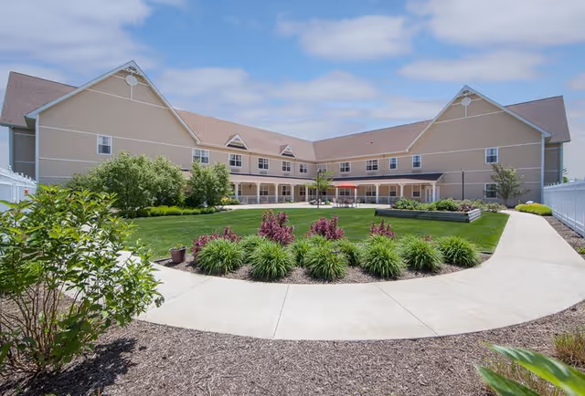 Outdoor courtyard area of a senior living facility with a large green lawn, landscaped plants, and a curved concrete walkway. The beige two-story building surrounds the courtyard with multiple windows and a covered patio area. The sky is partly cloudy.