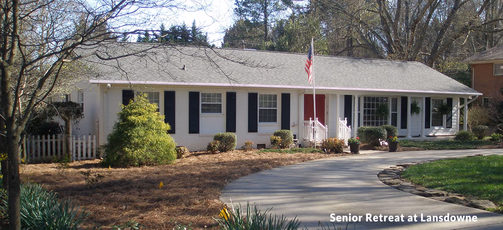Single-story white building with black shutters and a red door, surrounded by trees and shrubs, with a curved driveway leading to the entrance. An American flag is displayed on a flagpole near the front steps.