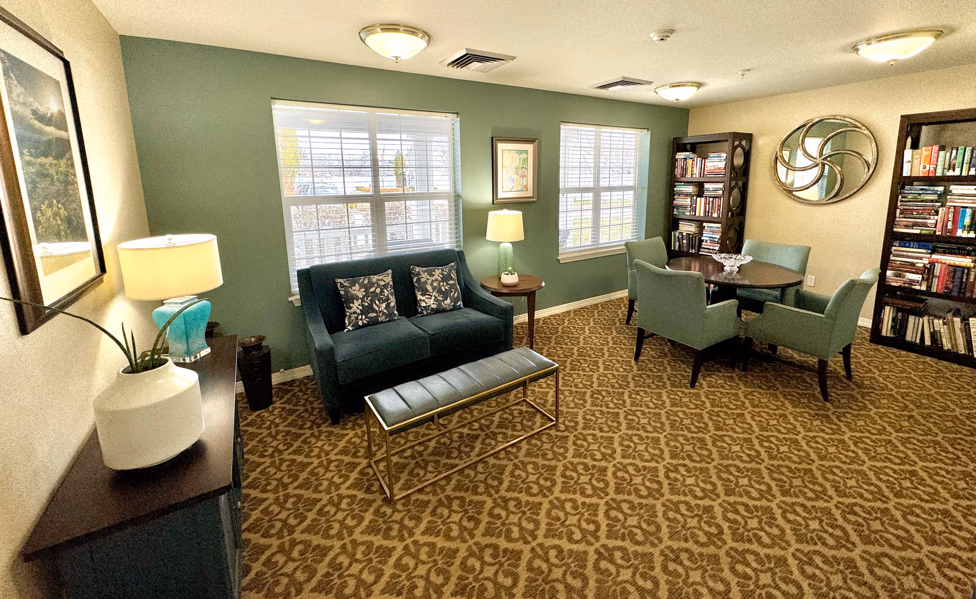 A cozy senior living common area with a patterned carpet, a dark green loveseat with two floral cushions, a small bench, a round table with four green upholstered chairs, two tall bookshelves filled with books, two windows with white blinds, a side table with a lamp, and a decorative mirror on the wall.