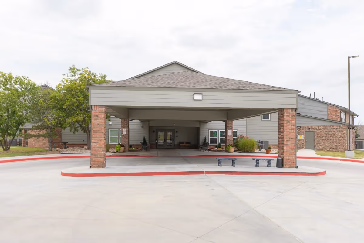Front view of a senior living facility entrance with a covered drive, brick columns, and landscaped plants.