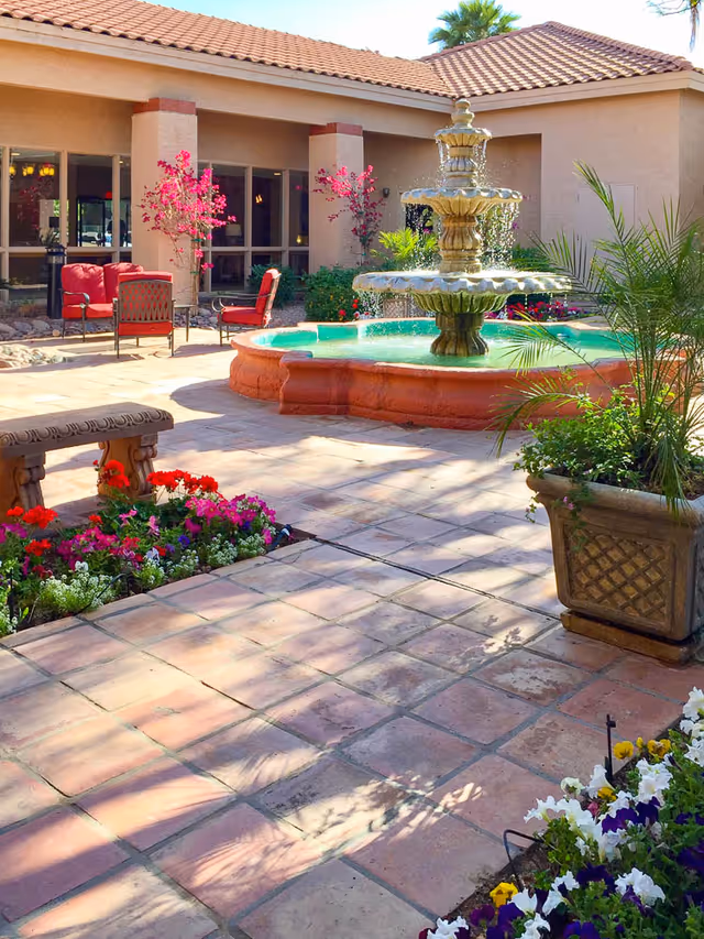 Sunny courtyard with a tiered fountain, potted plants, colorful flowers and patio chairs in front of a tile-roofed building.