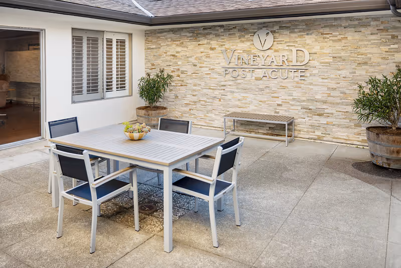 Outdoor patio area with a square table and four chairs, two large potted plants, a bench against a stone wall, and a sign on the wall that reads 'Vineyard Post Acute'.