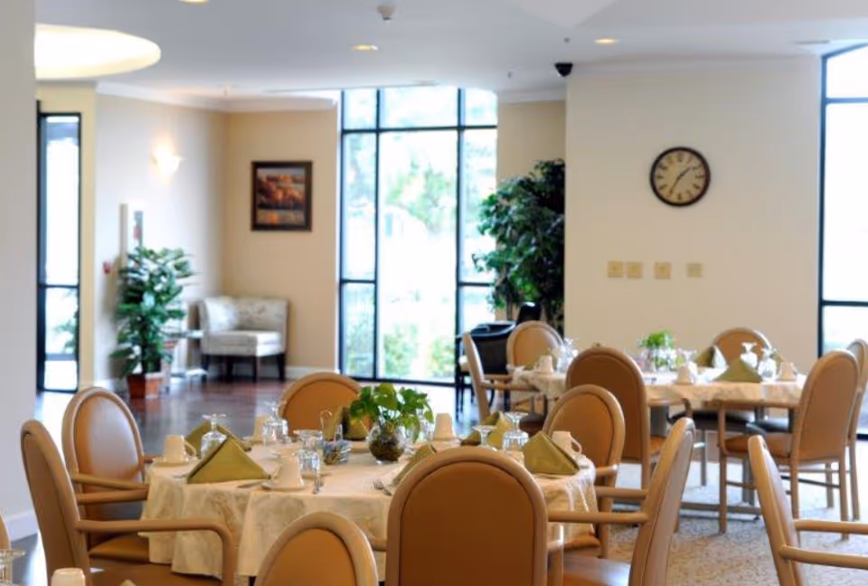 Well-lit dining room with round tables set with napkins and glassware, upholstered chairs, plants, and large windows.