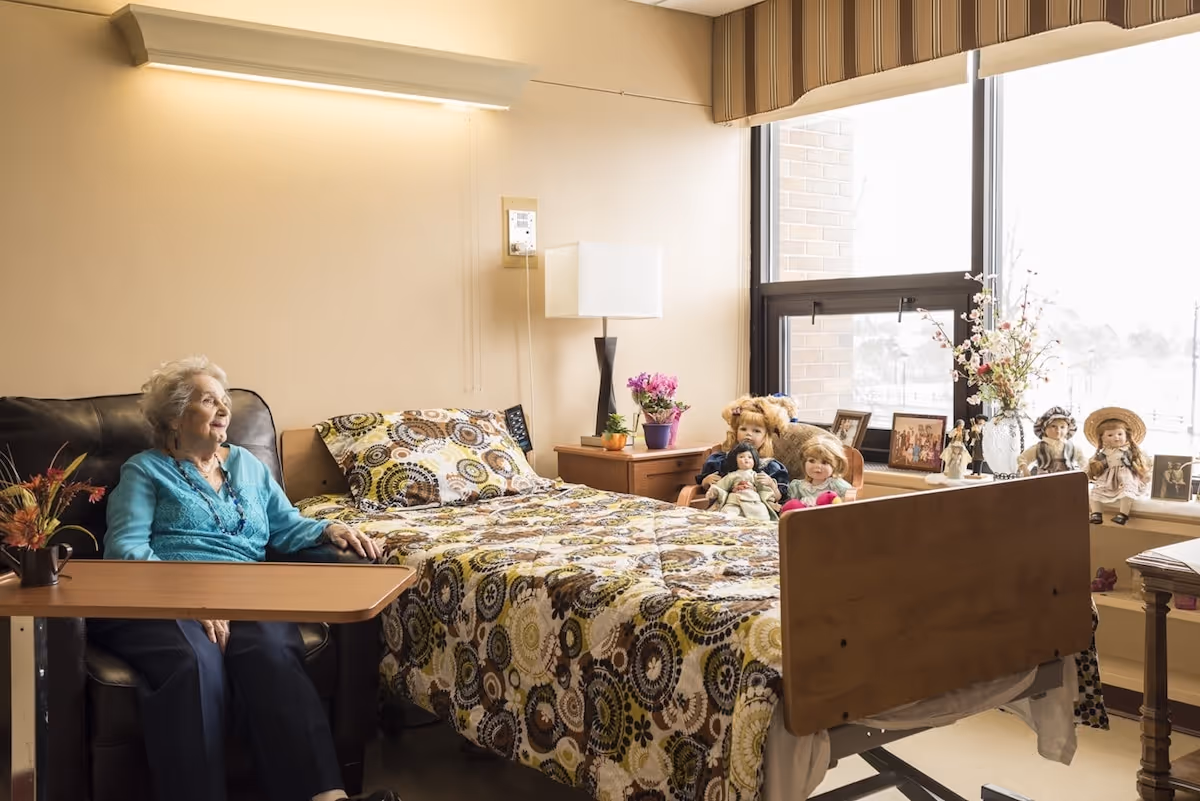 An elderly woman sitting on a black leather chair next to a hospital-style bed with a colorful patterned bedspread in a well-lit room. The room has a large window with a striped valance, a wooden nightstand with a lamp and potted plants, and several decorative dolls and framed photos on the windowsill.