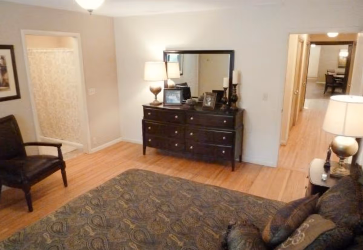 A bedroom with a patterned bedspread, a dark wooden dresser with a large mirror above it, two table lamps, framed photos, and candles. There is a black armchair to the left and a doorway leading to a bathroom with a shower curtain. Another doorway leads to a hallway with a view of a dining area in the distance.