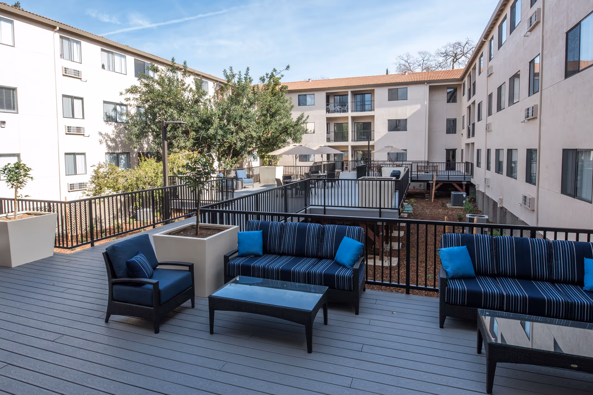 Outdoor patio area with blue cushioned chairs and sofas, glass-top coffee tables, potted plants, and a black metal railing. The patio is surrounded by a multi-story building with windows and air conditioning units visible. Trees and umbrellas are seen in the background under a clear blue sky.