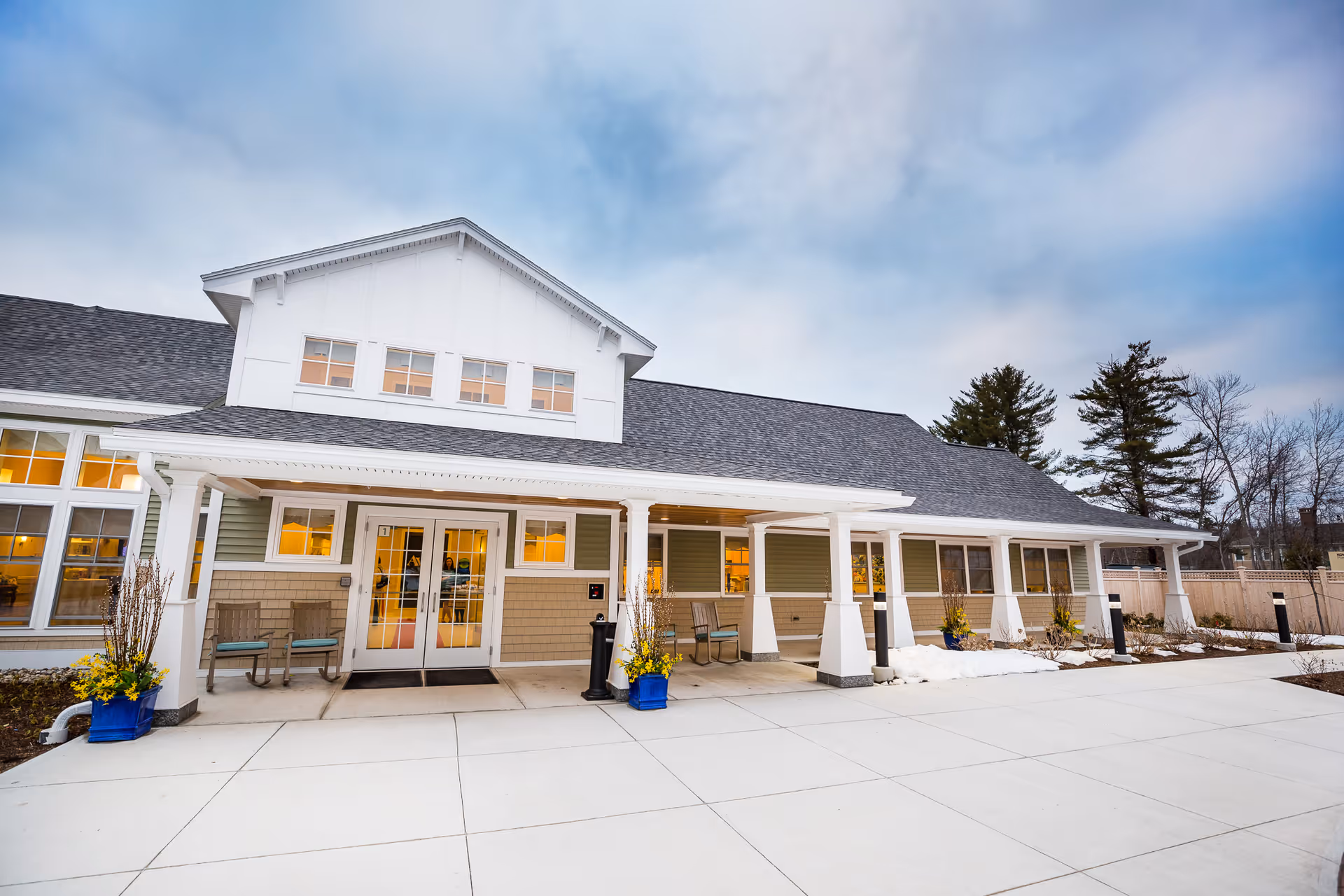 Exterior view of Avita of Wells senior living facility showing a single-story building with a covered entrance, large windows, and outdoor seating. There are planters with yellow flowers near the entrance and a paved walkway leading up to the doors. The sky is partly cloudy.