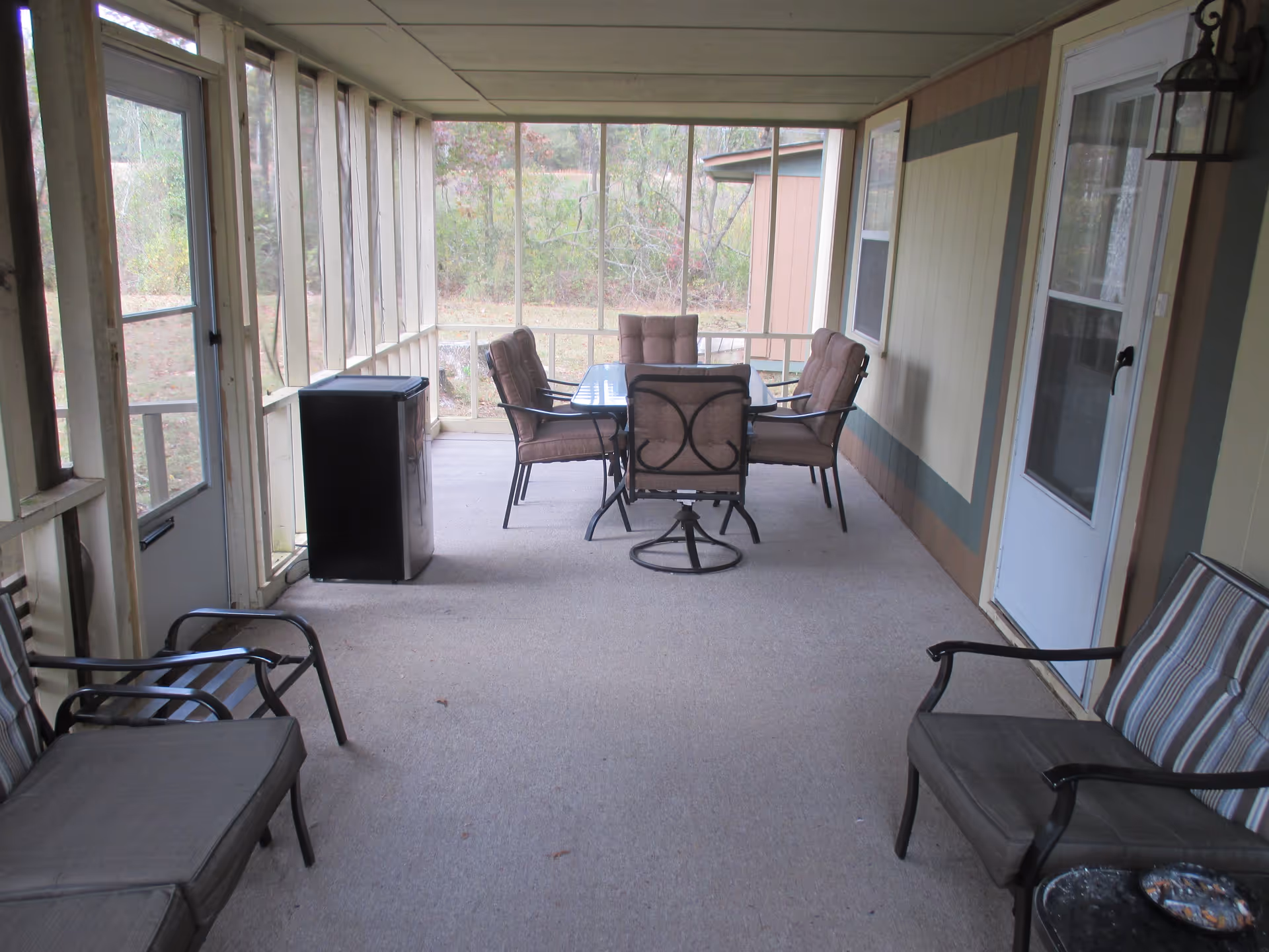 A screened-in porch with a glass-top table surrounded by six cushioned chairs in the center. Two cushioned armchairs and a cushioned loveseat are positioned along the sides. A small black refrigerator or cabinet is placed near the left side door. The porch overlooks a grassy outdoor area with trees.