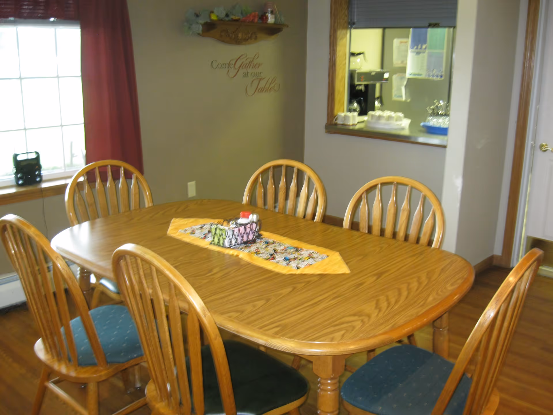 A wooden dining table with six wooden chairs around it, each chair having a cushioned seat in different colors. The table has a decorative runner with a small centerpiece. In the background, there is a window with red curtains and a wall with a decorative shelf and a sign that reads 'Come Gather at our Table'. There is also a pass-through window to a kitchen area with some items visible on the counter.