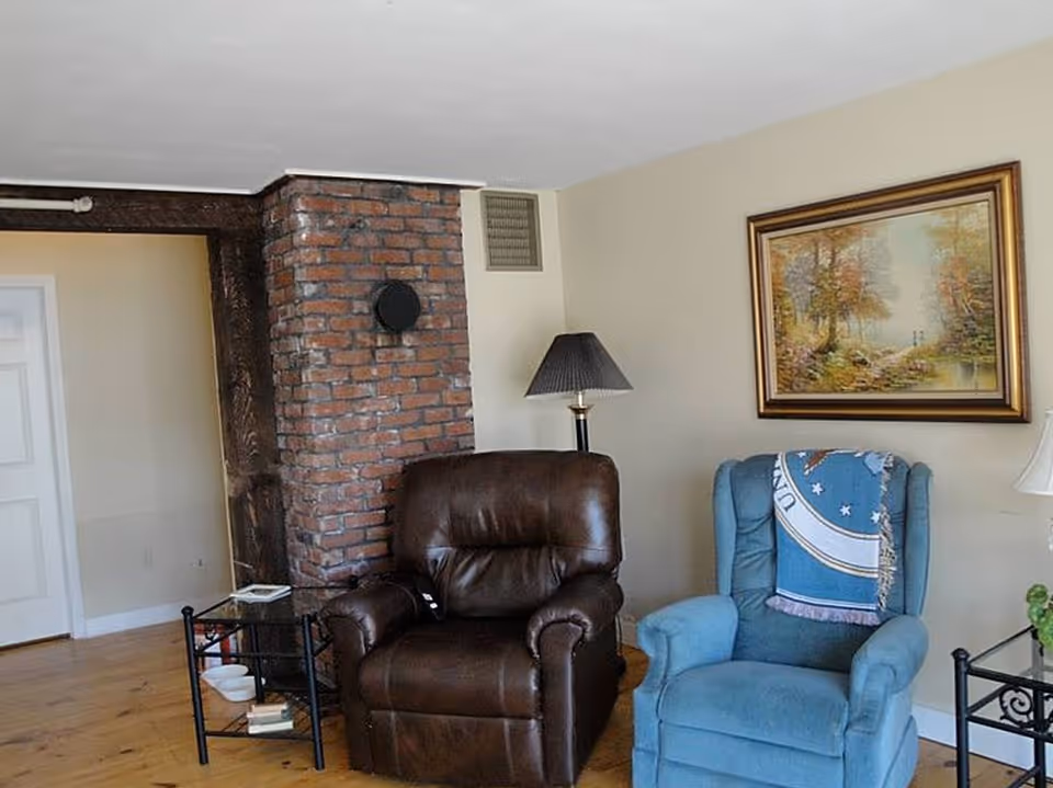 A cozy living room area featuring a brown leather recliner and a blue upholstered armchair with a decorative throw draped over the back. Between the chairs is a small black metal side table with glass shelves holding some books and a white bowl. Behind the brown recliner is a brick chimney, and on the wall above the blue chair hangs a framed painting of a serene forest scene with a river. A floor lamp with a dark shade stands between the chairs, and the room has light-colored walls and wooden flooring.
