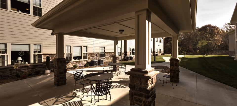 Outdoor covered patio area with round metal tables and chairs, surrounded by a paved walkway and green lawn, adjacent to a beige building with multiple windows.