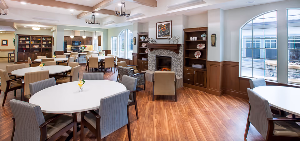 A spacious and well-lit dining area with multiple round tables surrounded by chairs. The room features large arched windows letting in natural light, a fireplace with decorative shelves on either side, and a kitchen area in the background. The floor is wooden, and the walls are painted in light colors with brown wainscoting.
