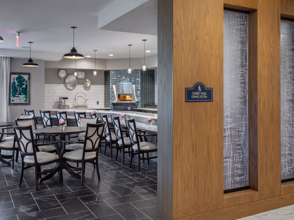 Dining room with multiple round tables and chairs, a counter and open kitchen with a pizza oven, and a wooden partition labeled 'Torrey Pines Dining Room'.