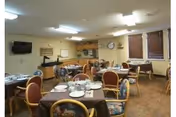 A dining room in a senior living community with multiple round tables covered with brown tablecloths, set with plates, bowls, and utensils. The room has cushioned chairs with floral upholstery, a wall-mounted television, a clock, and windows with wooden blinds. The lighting is provided by ceiling fixtures.